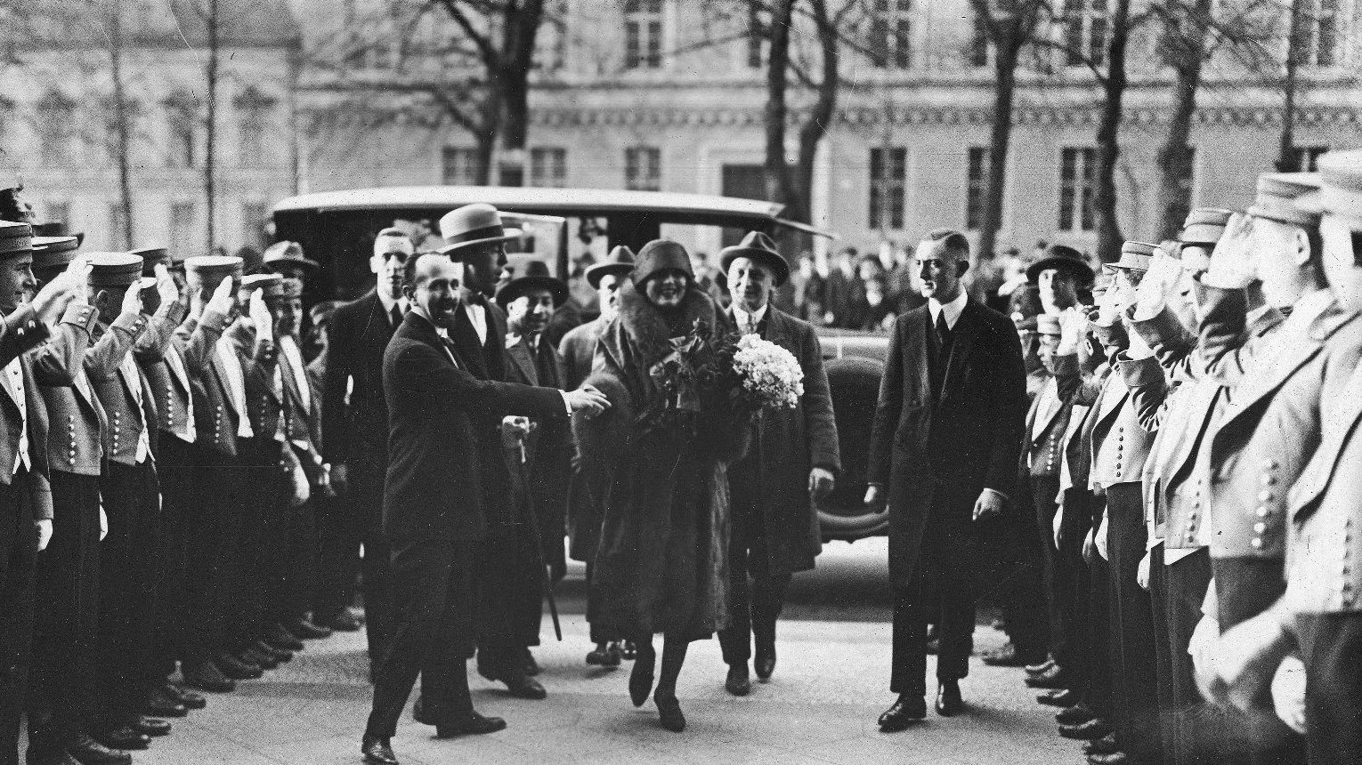 Kobieta w futrze wychodzi z samochodu w towarzystwie kilku mężczyzna. Po prawej i lewej stronie rząd osób z podniesionymi rękami.