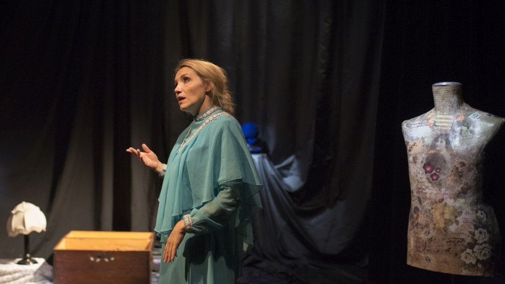 Aktorka deklamuje monolog na scenie. Za nią rekwizyty: manekin, lampka nocna, drewniana skrzynka.
