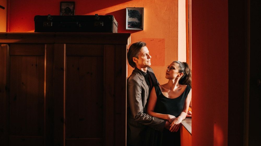 Obejmująca się para stoi przy oknie obok starej szafy w pokoju o pomarańczowych ścianach.