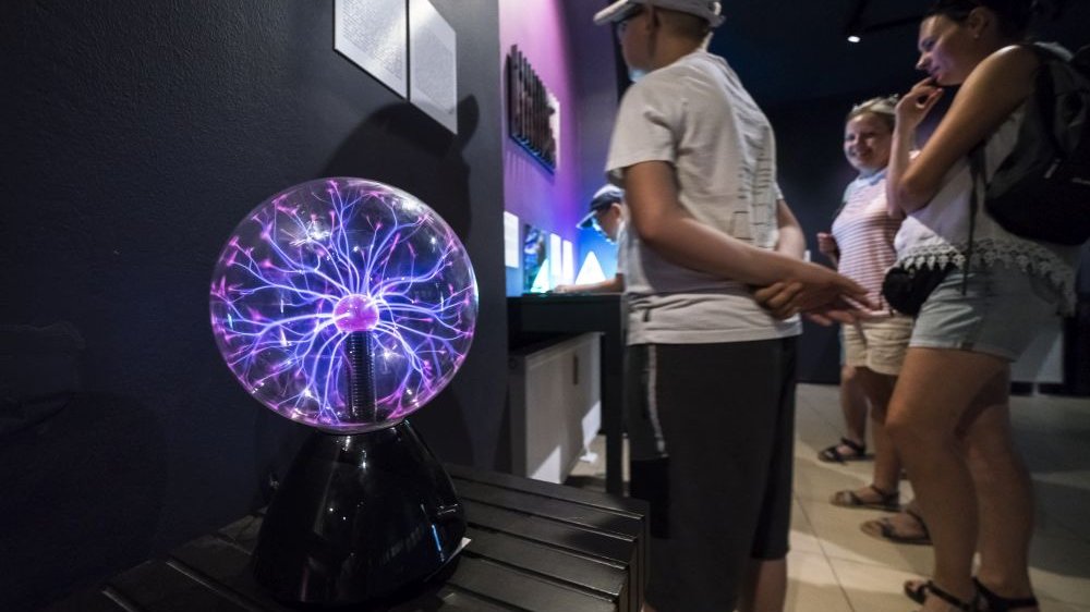 Zwiedzający muzem przyglądają się eksponatom, na zdjęciu widać wyraźnie jeden z nich - szklaną kulę, w której buzują fioletowe i różowe promienie światła.