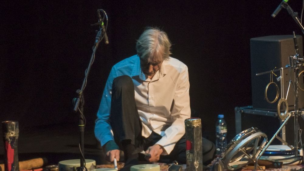 Mężczyzna w białej koszuli siedzi na ziemi, dookoła niego drobne instrumenty perkusyjne.