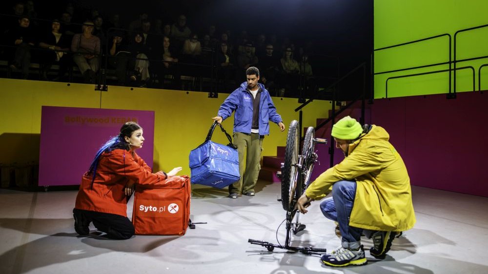 Trojka aktorów w kolorowych kurtkach na scenie. Jeden z nich naprawia rower, reszta trzyma duże torby do przewożenia jedzenia.