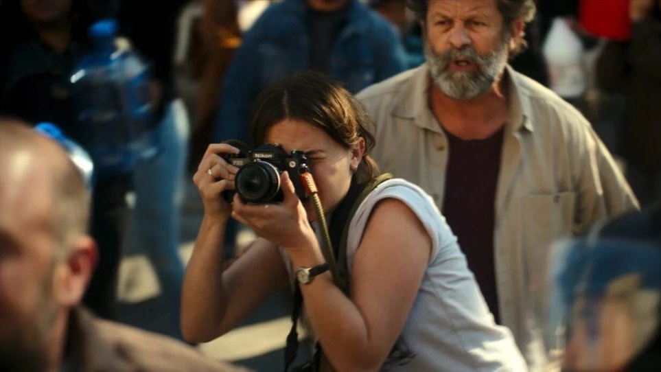 Kobieta fotografuje coś w tłumie ludzi. Przyjęła wygiętą pozycję, aparat trzyma tuż przy swojej twarzy.