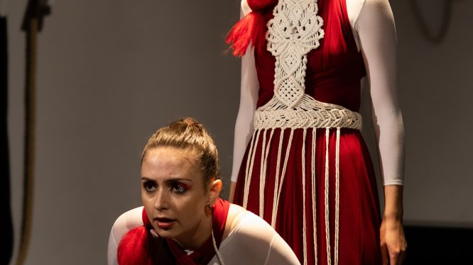 Dwie aktorki w biało-czerwonych strojach z elementami ludowych plecionek. Jedna kuca, druga stoi za jej plecami.
