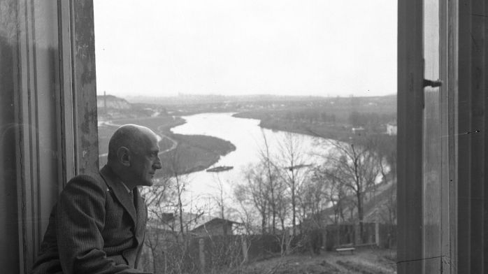 Łysy mężczyzna w garniturze siedzi przy otwartym oknie, patrzy w dal. Za oknem widoczna szeroka rzeka, pola i drzewa.