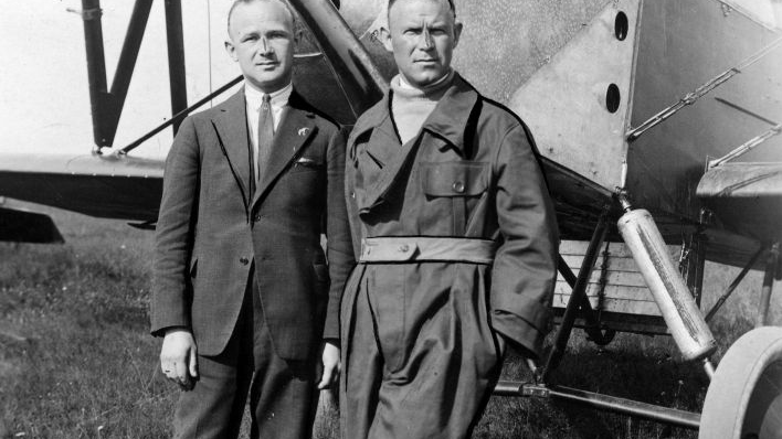 Dwaj mężczyźni, jeden w garniturze, drugi w kombinezonie lotniczym, przed dziobem samolotu