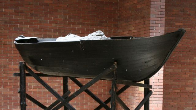 Czarna łódka stoi na drewnianym stelażu. Z tyłu ceglane ściany.