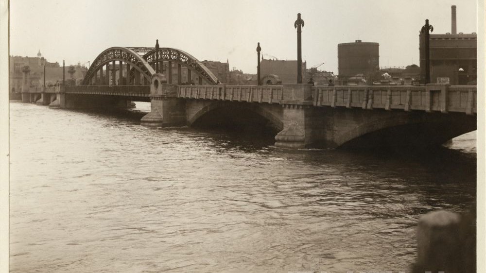 Zdjęcie mostu z dwoma półokrągłymi przęsłami. W tle budynki.