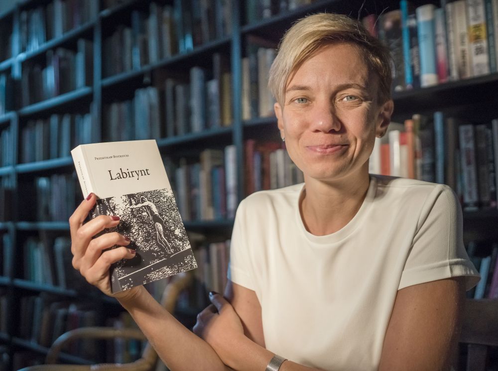 Uśmiechnięta kobieta trzyma w ręku książkę pt."Labirynt", siedzi. Za jej plecami półki pełne książek. - grafika artykułu