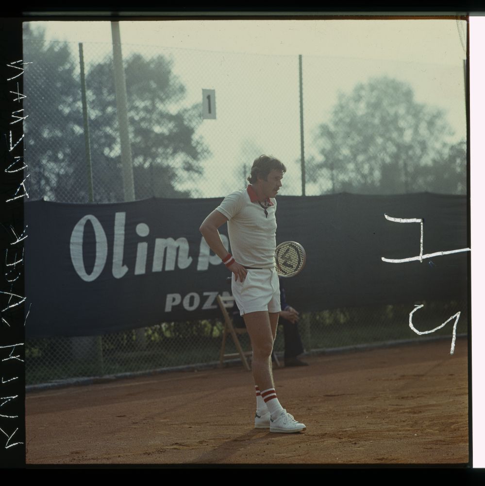 Meżczyzna z wąsem w stroju tenisowym stoi na korcie, ręce opiera na biodrach. - grafika artykułu