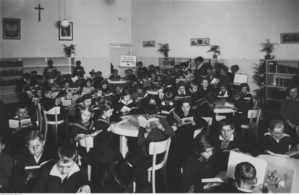 Czarno-białe zdjęcie dużej grupy dzieci w mundurkach szkolnych. Siedzą przy stolikach, czytają książki. - grafika artykułu