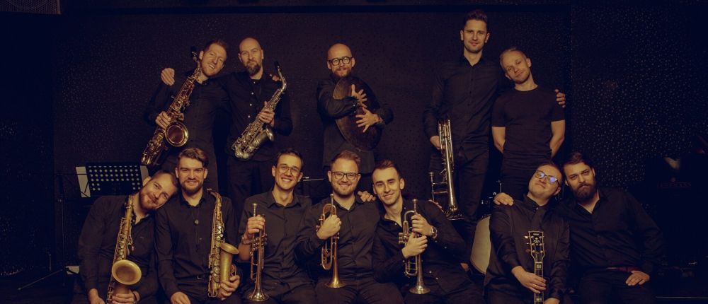 Grupowe zdjęcie dwunastu muzyków orkiestry. Mężczyźni są ubrani w ciemne koszule, każdy z nich trzyma w ręce instrument. - grafika artykułu