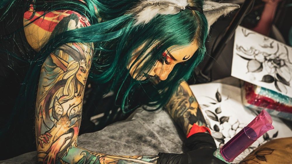Wytatuowana dziewczyna z zielonymi włosami i w mocnym makijażu tatuuje klienta.