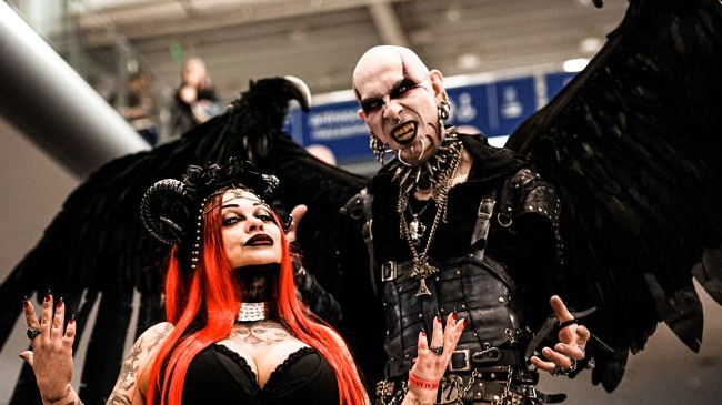 Para w gotyckich strojach stoi prezentując swoje outfity. Kobieta ma długie, czerwone włosy i czarne rogi na głowie, mężczyzna dużo kolczyków i makijaż przypominający postać filmowego Nosferatu.