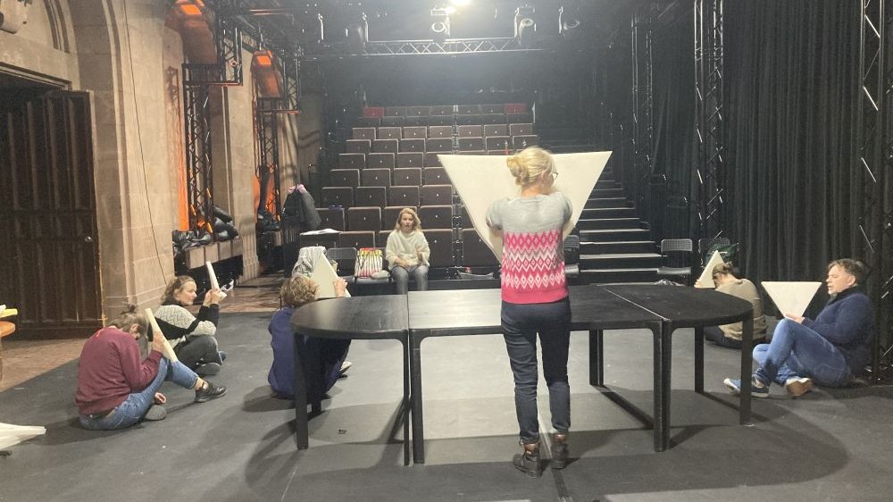 Zespół teatru próbuje spektakl na scenie. Na środku stoi kobieta z odwróconym, piankowym trójkątem w rękach.
