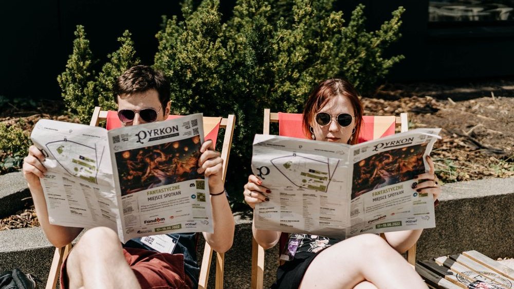 Chłopak i dziewczyna, oboje w przeciwsłonecznych okularach, siedzą na leżakach czytając gazetę festiwalową.