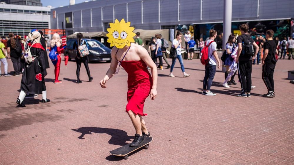 Uczestnik przebrany za postać Lisy Simpson z serialu animowanego "Simpsonowie" jedzie na deskorolce. Za nim uczestnicy Pyrkonu na dziedzińcu MTP.