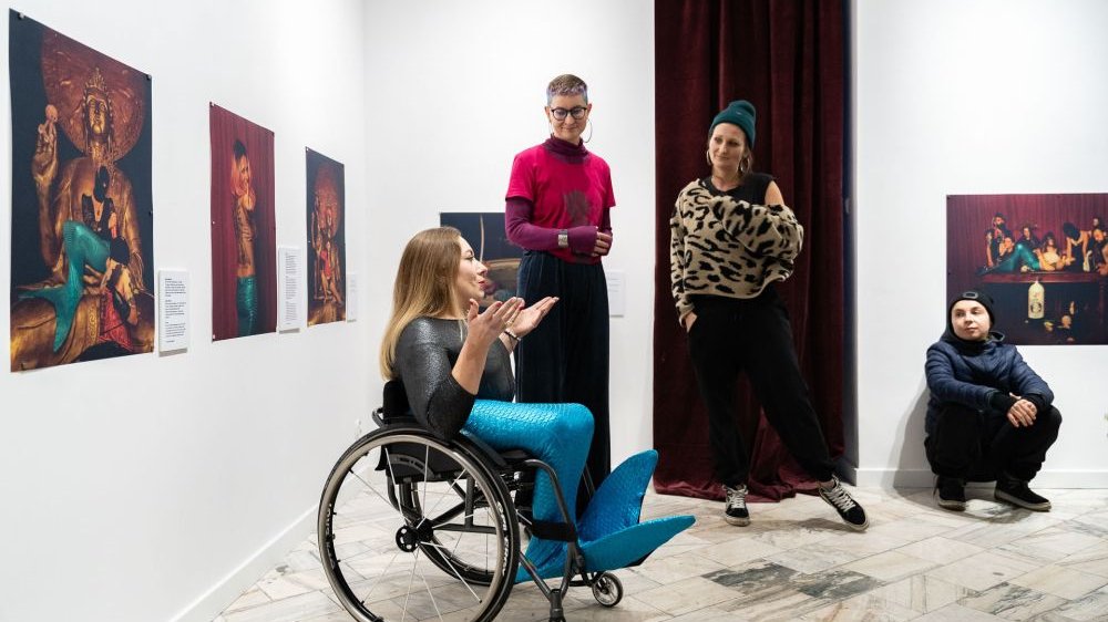 Na ścianach wiszą zdjęcia w nasyconych, czerwonych i fioletowych kolorach. Przed ekspozycją stoją artystki i kuratorki, jedna z nich jest osobą z niepełnosprawnością na wózku inwalidzkim i ma na sobie błękitny, syreni ogon zamiast nóg.
