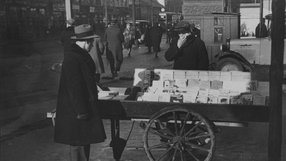 Czarno-białe zdjęcie ulicznego sprzedawcy książek. Jego stoisku przygląda się drugi mężczyzna, w tle inni przechodnie.